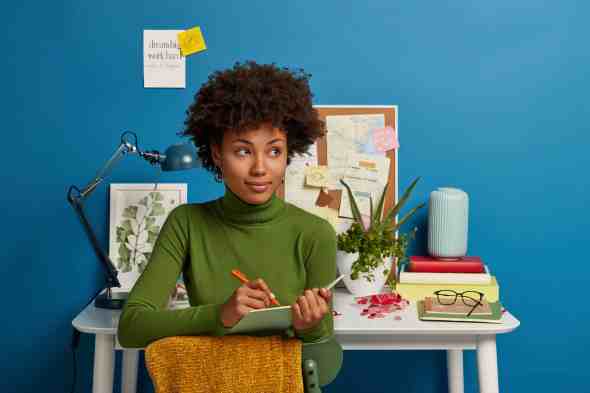Pensive curly young girl writes down future plans and goals in notepad, thinks about good idea, poses against cozy workplace interior, dressed in comfortable green jumper, isolated on blue wall